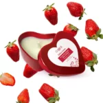 Une bougie rouge en forme de cœur, avec un couvercle partiellement ouvert et des fraises délicatement disposées autour.